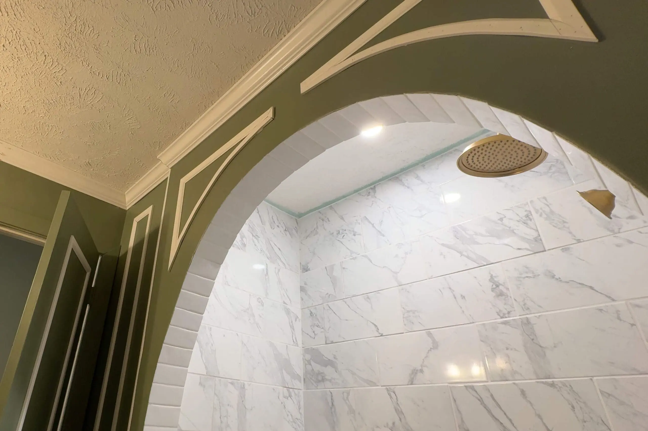 Elegant shower archway with green walls featuring white curved trim. The shower interior showcases glossy white marble-style tiles with a recessed shelf and a modern gold rain showerhead, highlighted by recessed lighting above.